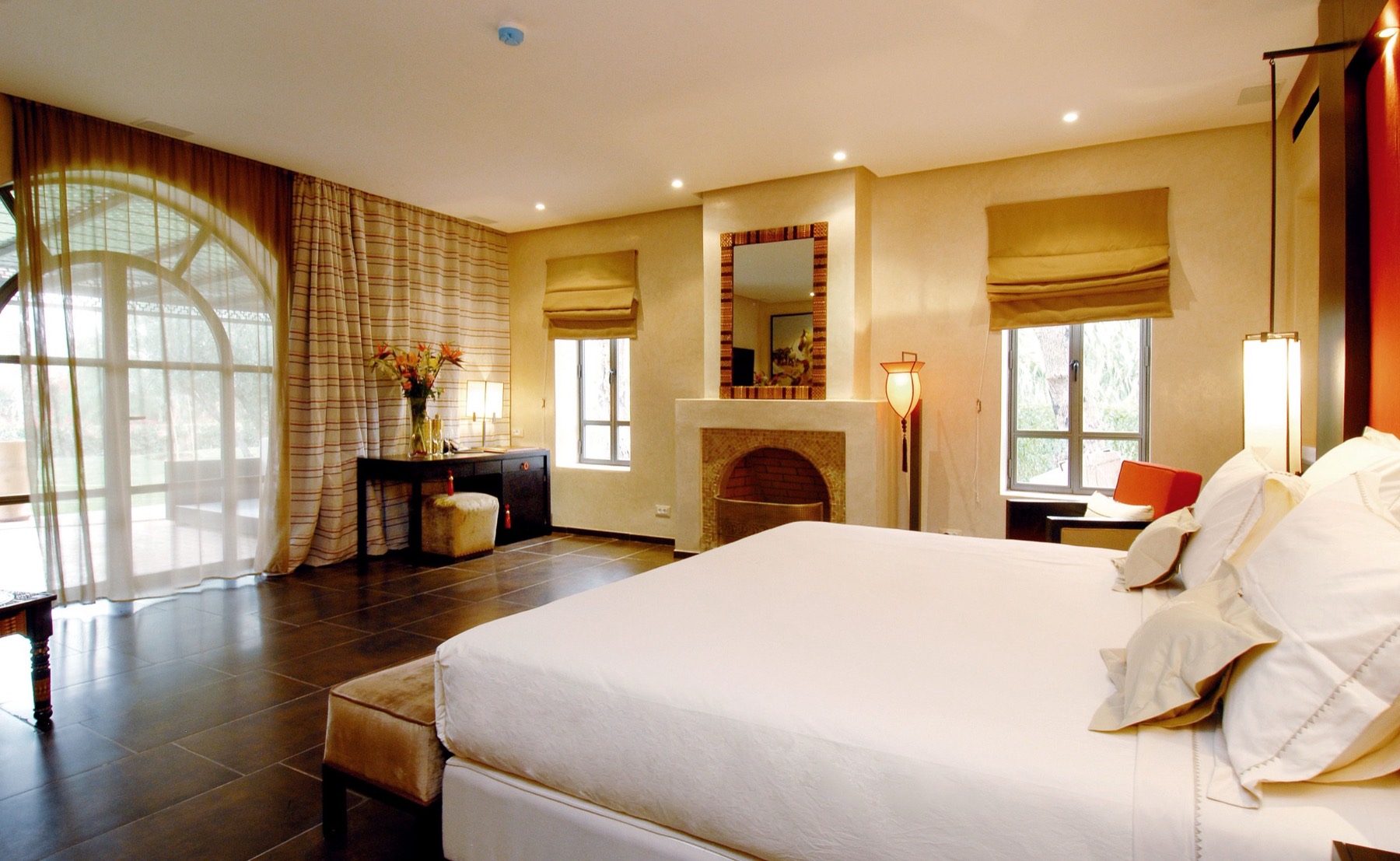 Warm sunlit boutique hotel suite with limewashed clay walls, a large arched window, a low king bed and dark terracotta tile floor