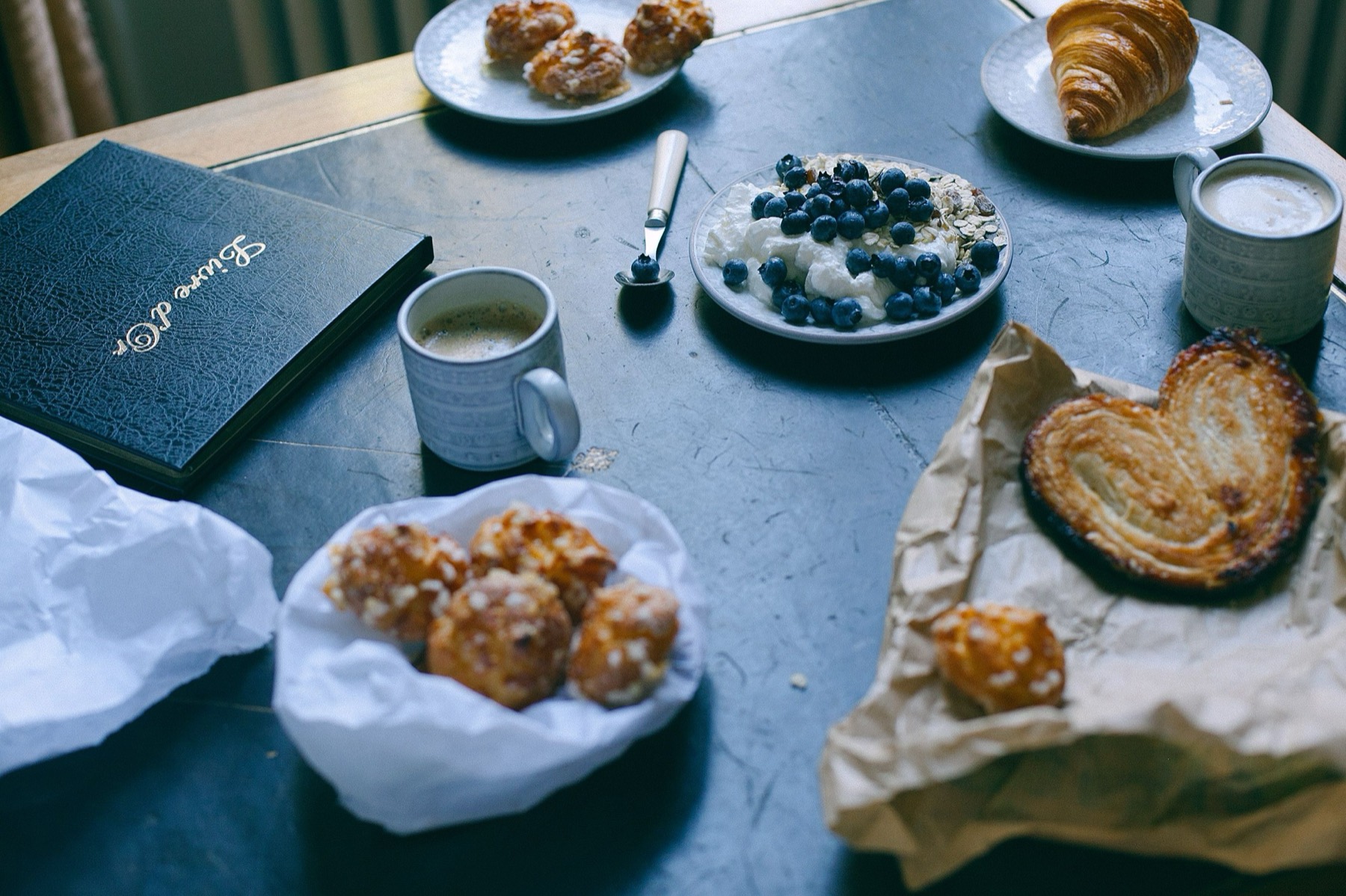 Overhead breakfast spread with pastries, a palmier, blueberries and yoghurt, coffee and a leather guestbook on a dark table
