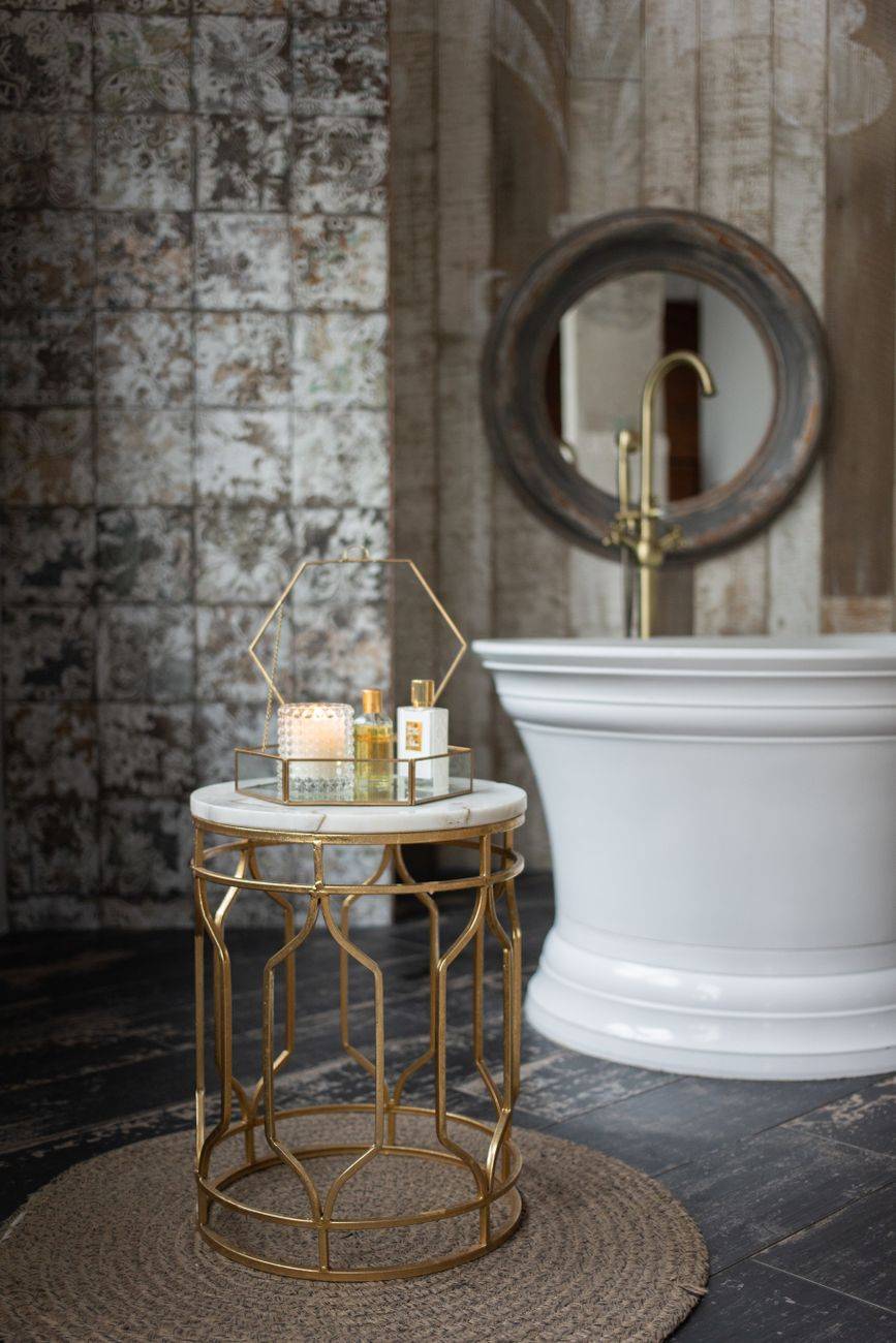 Glass box with spa supplies placed on small round table near white bathtub in stylish bathroom near wall with mirror