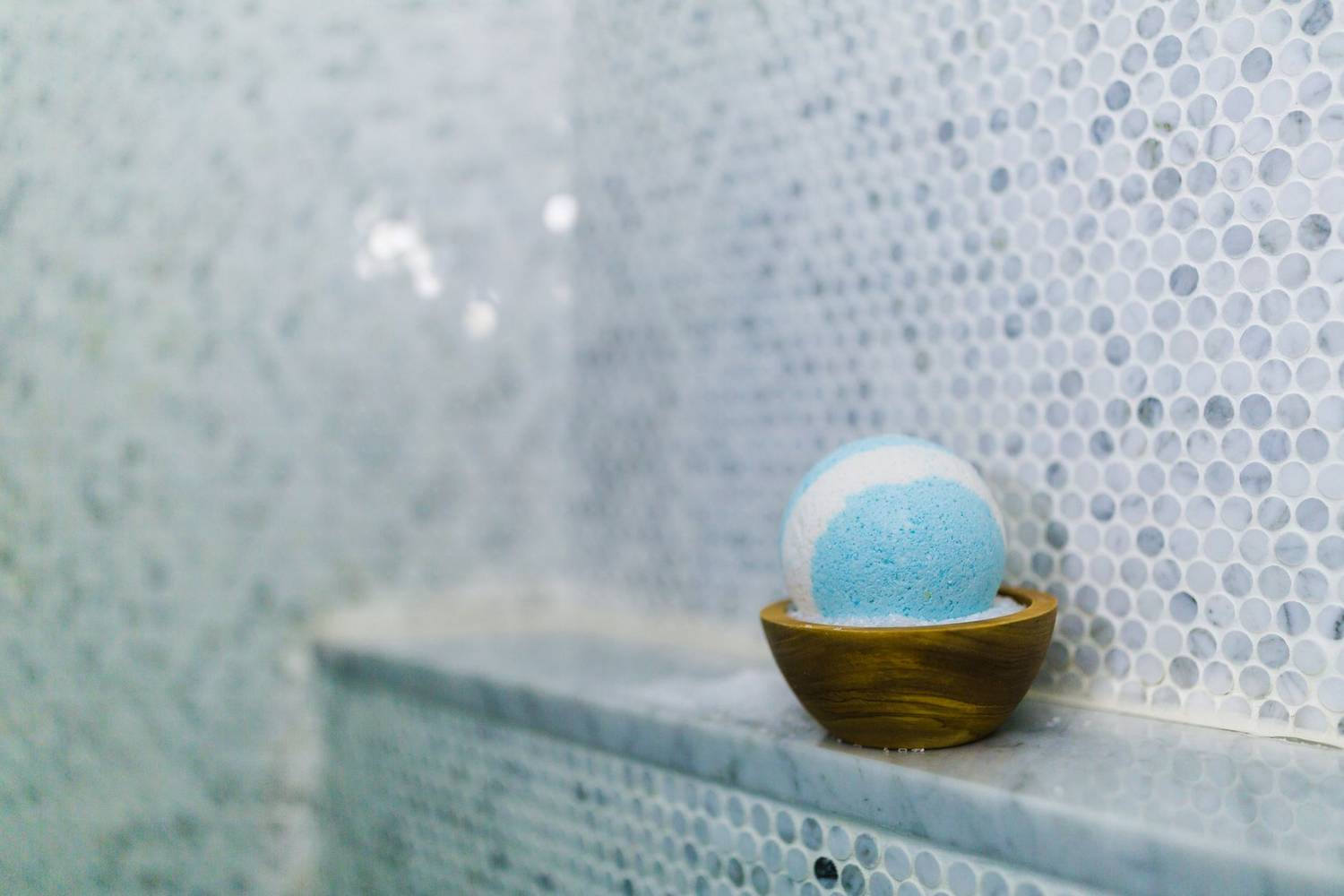Blue bath bomb in wooden bowl on marble bathroom shelf with stylish tiles.
