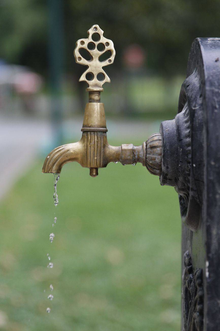 A close-up of a vintage brass faucet with water dripping, set in a park outdoor setting.
