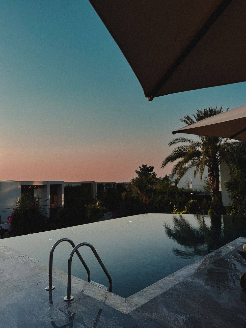 A tranquil infinity pool reflecting the colors of a stunning sunset, surrounded by palm trees.