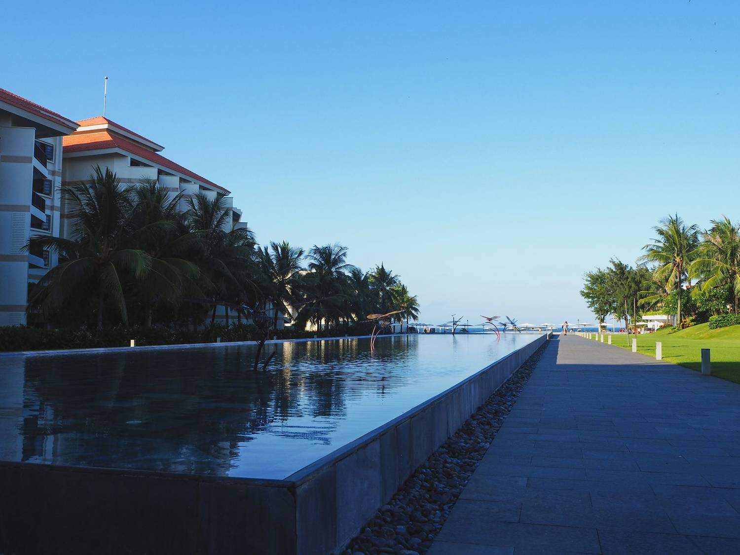 Exterior of modern tropical hotel with swimming pool and lush palm trees in yard located at seaside against cloudless blue sky in sunlight
