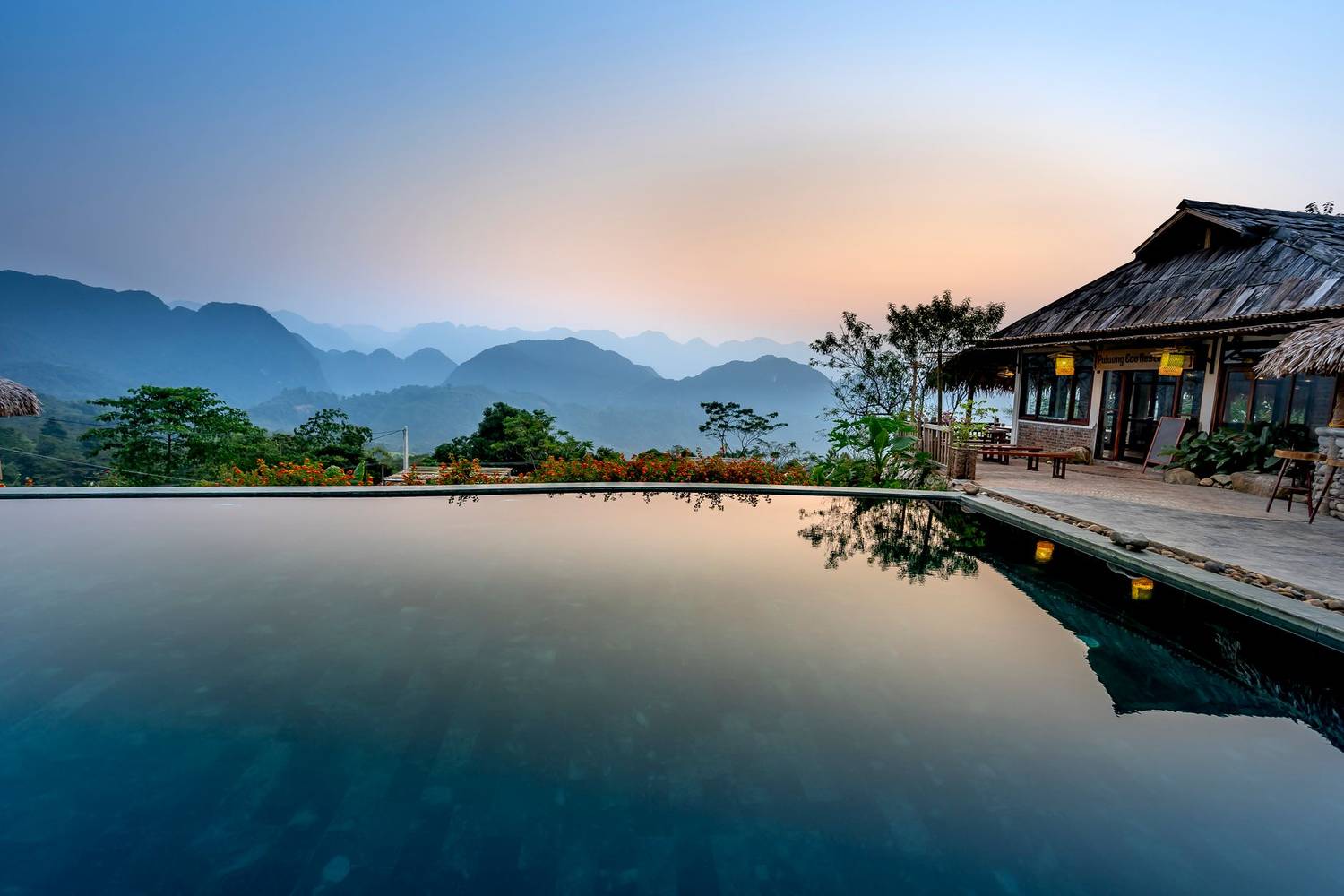 Pool with calm water located near terrace of tropical resort with thatched house against mountain ridge and green trees in nature