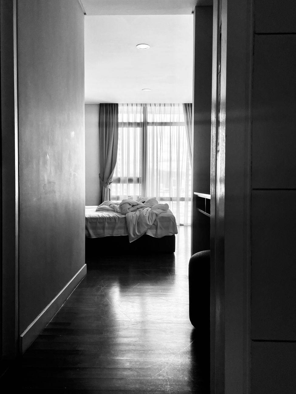 Black and white image of an elegant bedroom with unmade bed and soft sunlight through curtains.