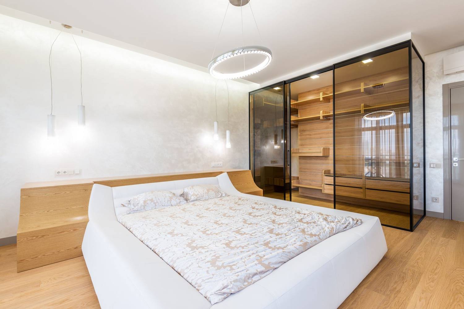 Modern interior design of cozy luxury bedroom with cozy king sized bed and glass wardrobe closet lighted with led chandelier and bedside lam