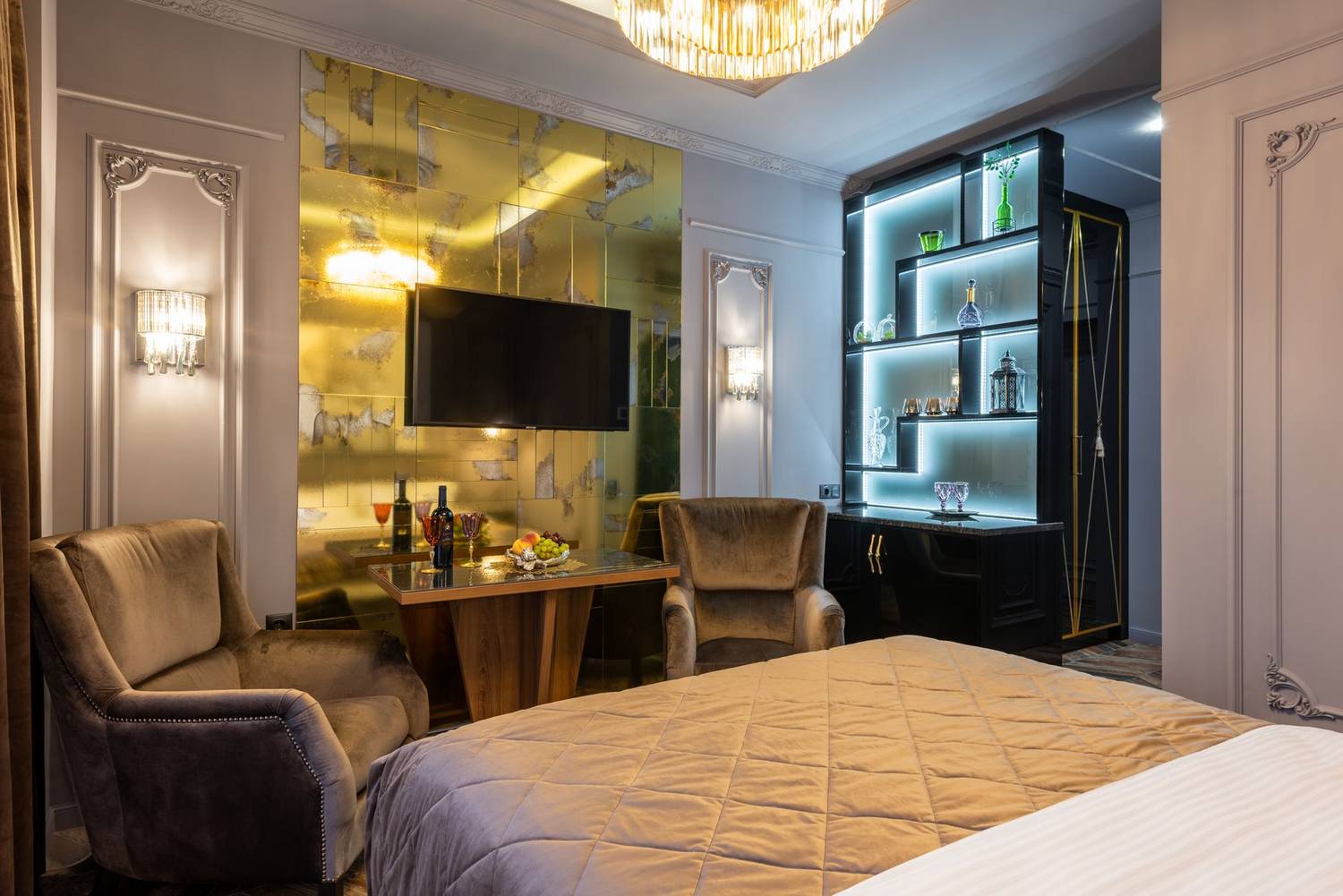 LCD television set against bed with cover in house with table between armchairs and shiny chandelier