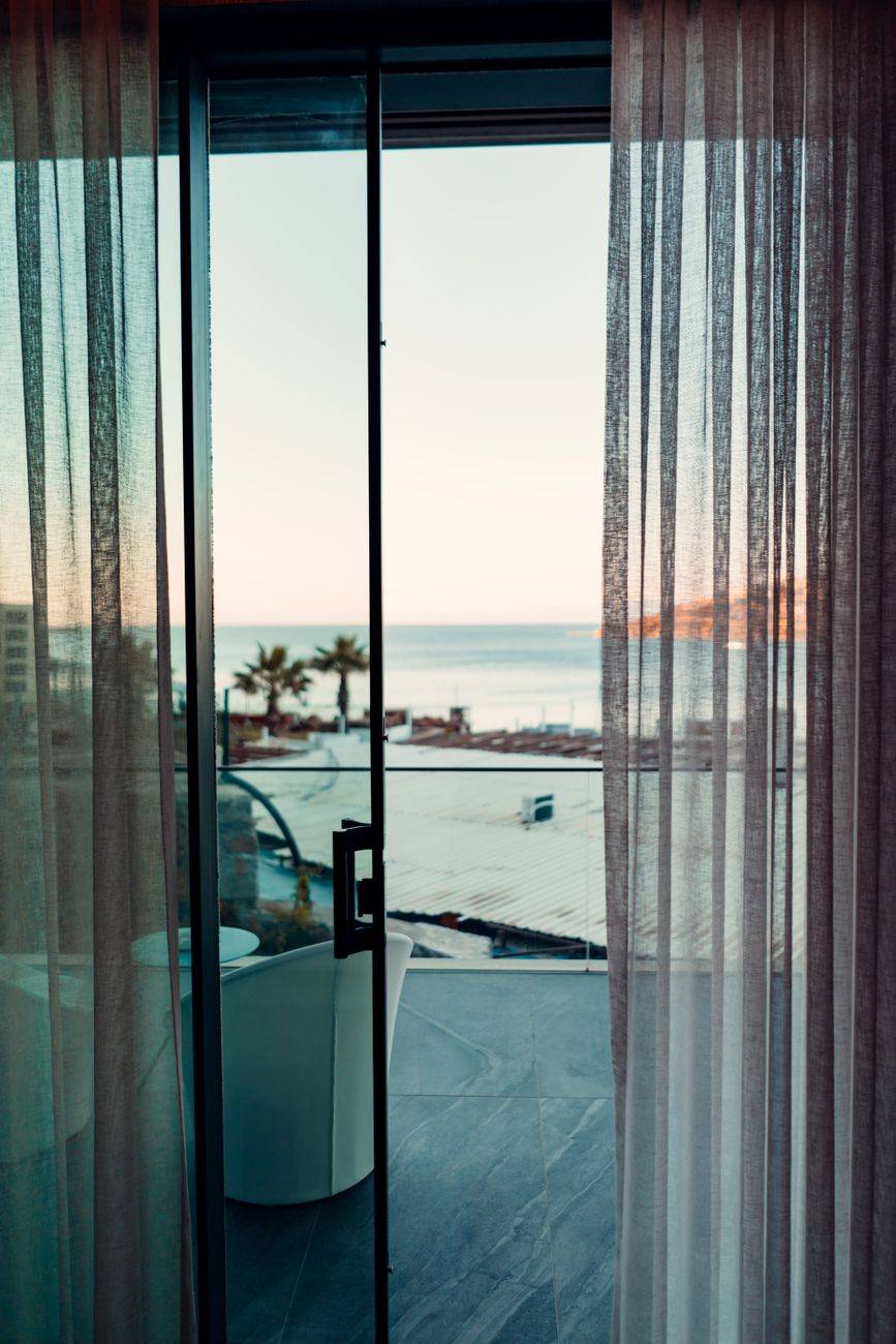 A tranquil ocean view through sheer curtains from a hotel room balcony at sunset.