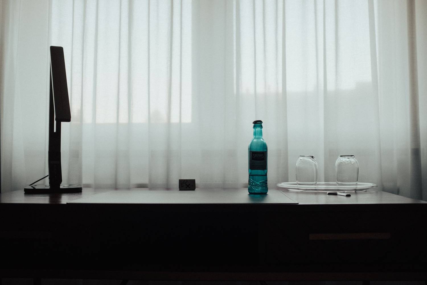 Simple desk with a blue bottle and empty glasses beside a soft lit window.