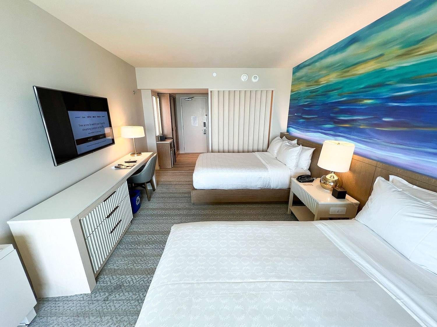 Spacious hotel room in Honolulu featuring two beds, vibrant ocean artwork, and modern amenities.