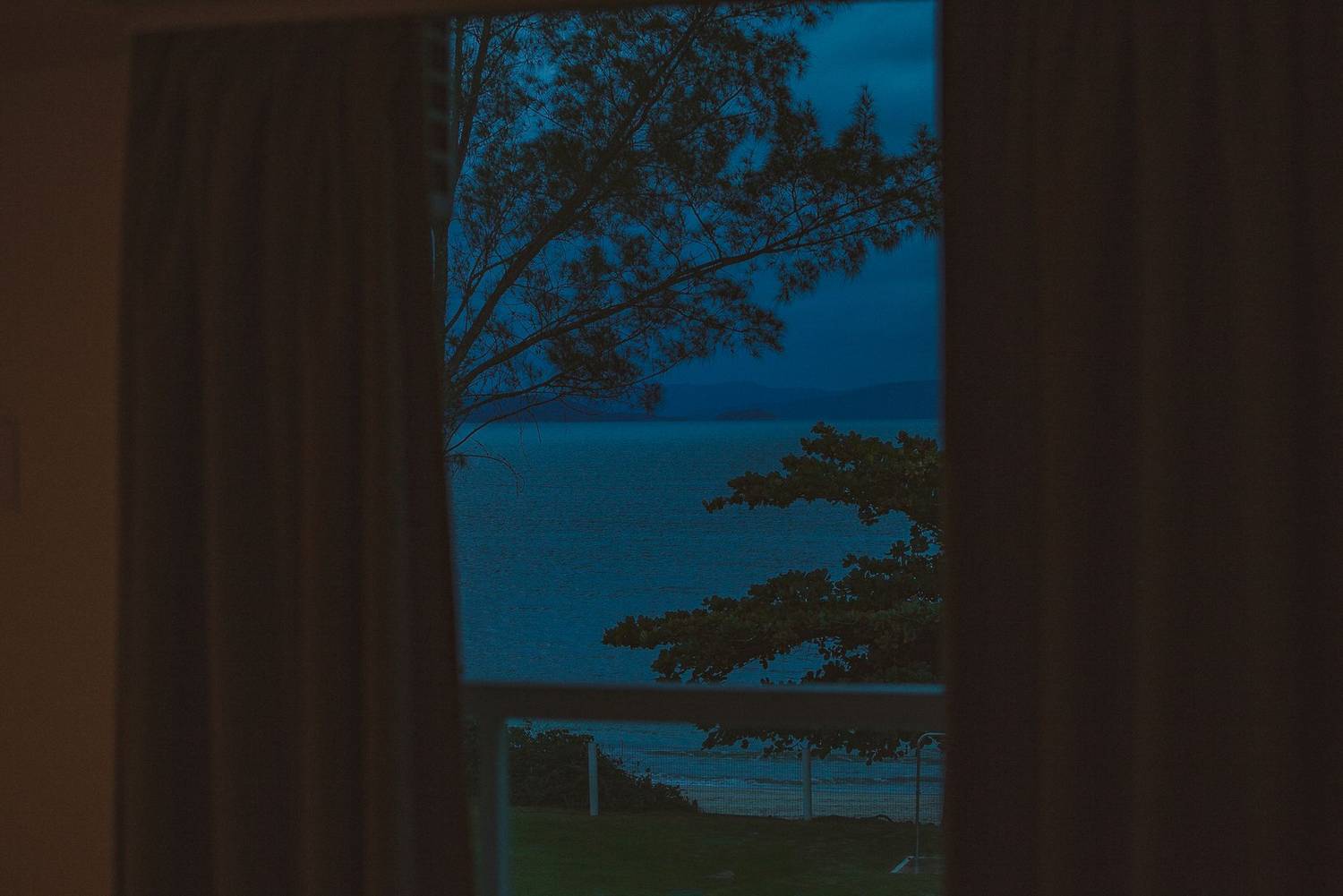 From fenced balcony view of silent ocean near trees behind mount silhouettes under sky at dusk