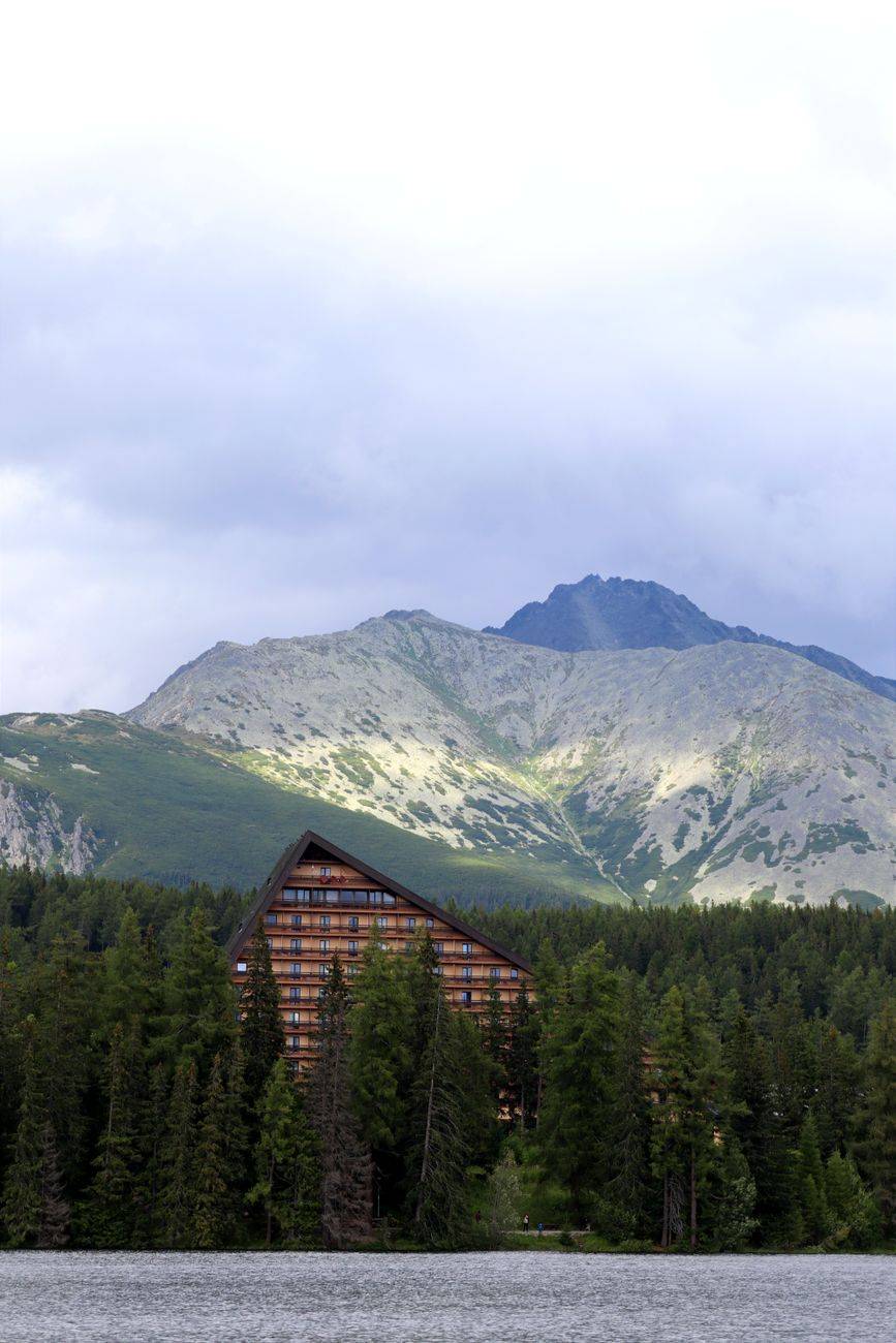 A serene mountain lodge nestled in lush forest by a tranquil alpine lake, under a cloudy sky.