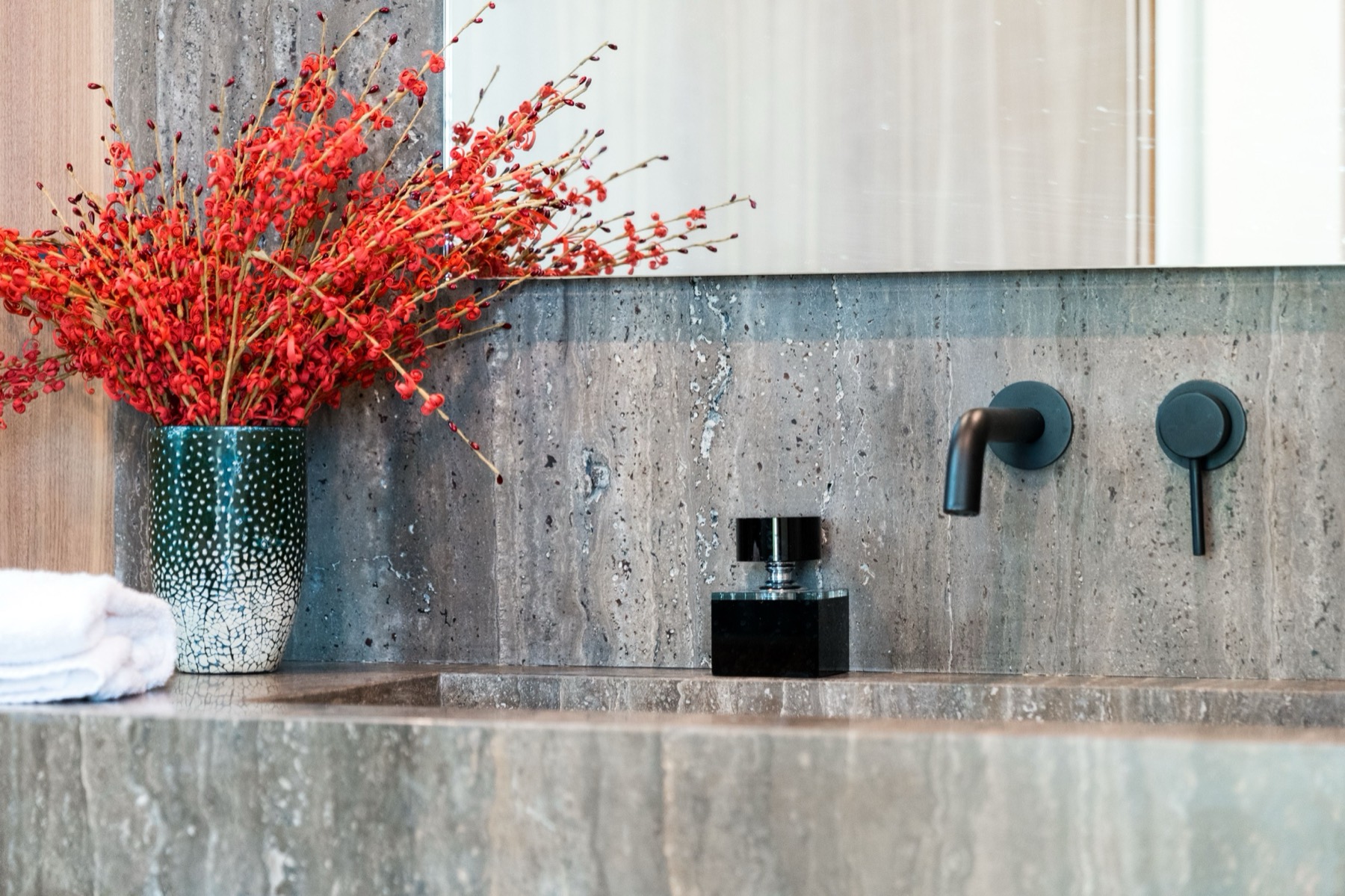 An interior styling detail of a travertine vanity with a folded towel and a vase of red branches