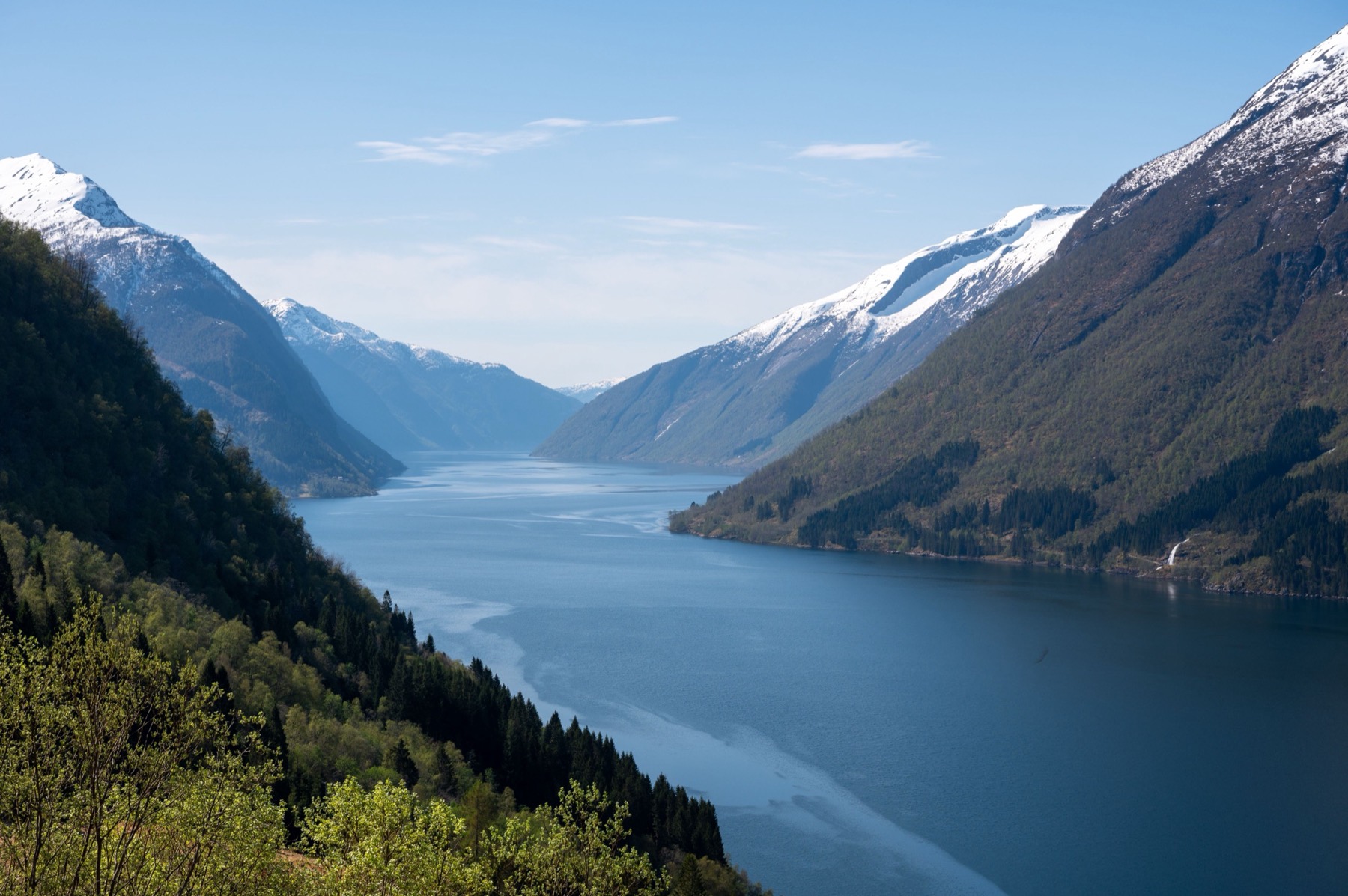 A blue Norwegian fjord flanked by steep green slopes and snow-streaked peaks under clear sky
