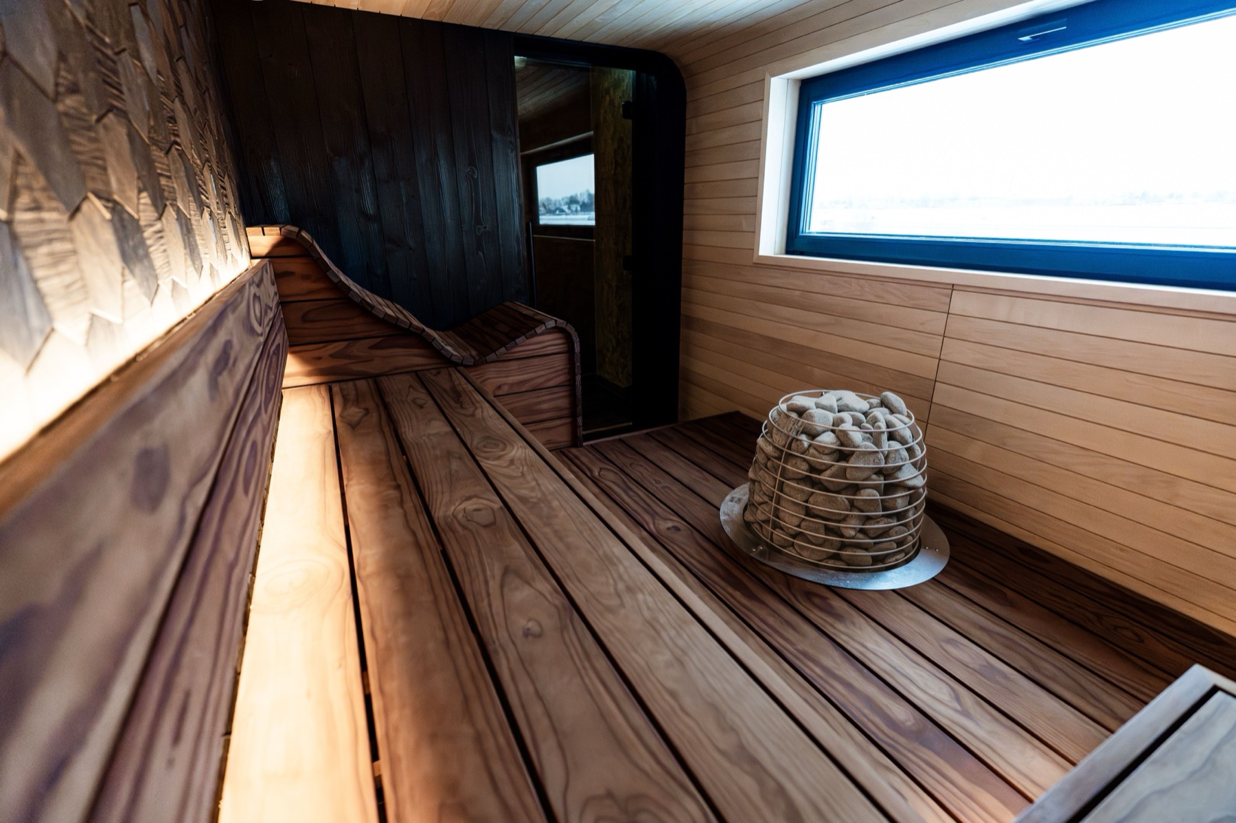 Wood-lined sauna interior with slatted benches, a stone-filled heater and a panoramic window onto water